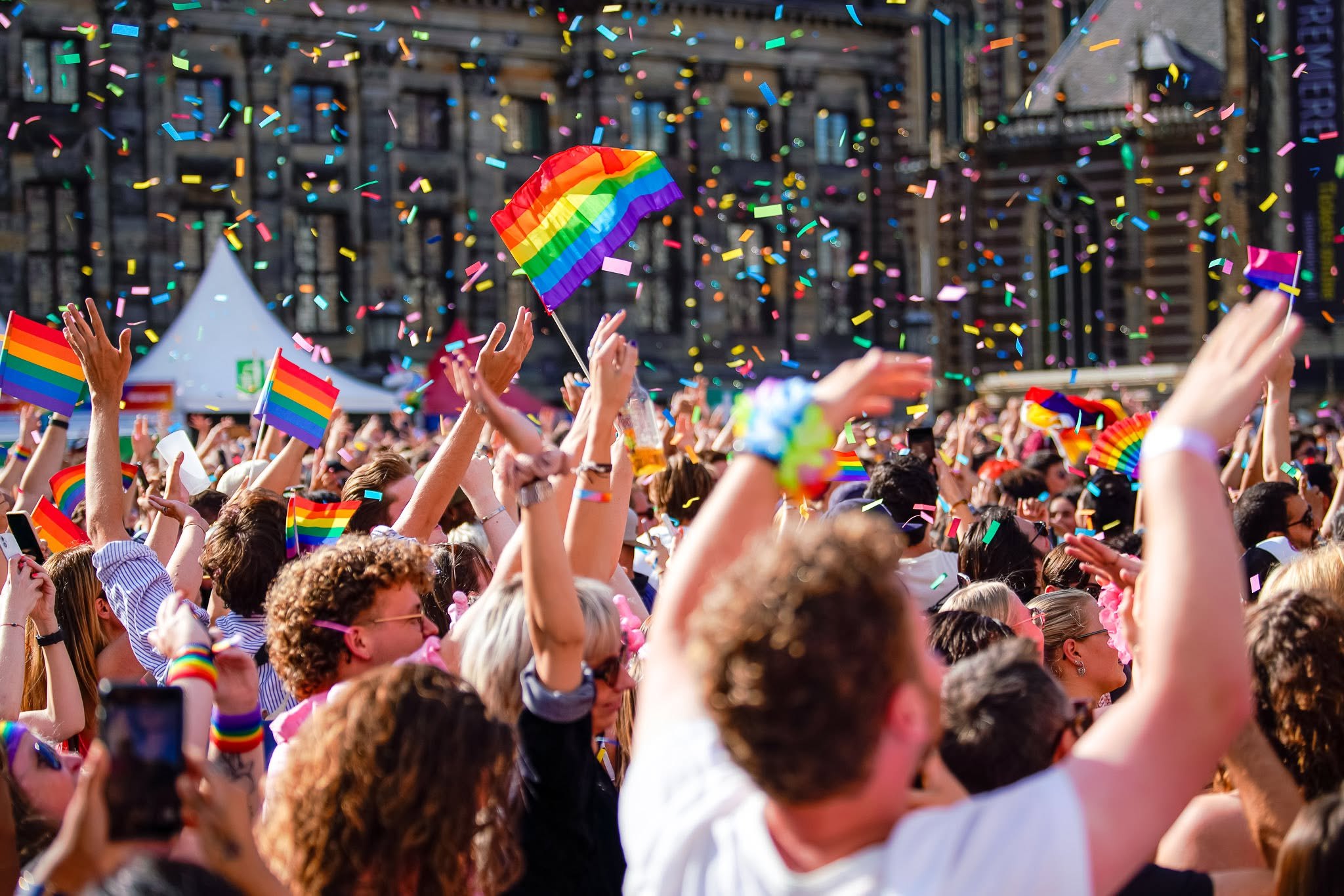 Het publiek bij MainStage Pride Amsterdam op de Dam, armen omhoog, regenboogvlaggen en confetti in de lucht voor het Koninklijk Paleis.