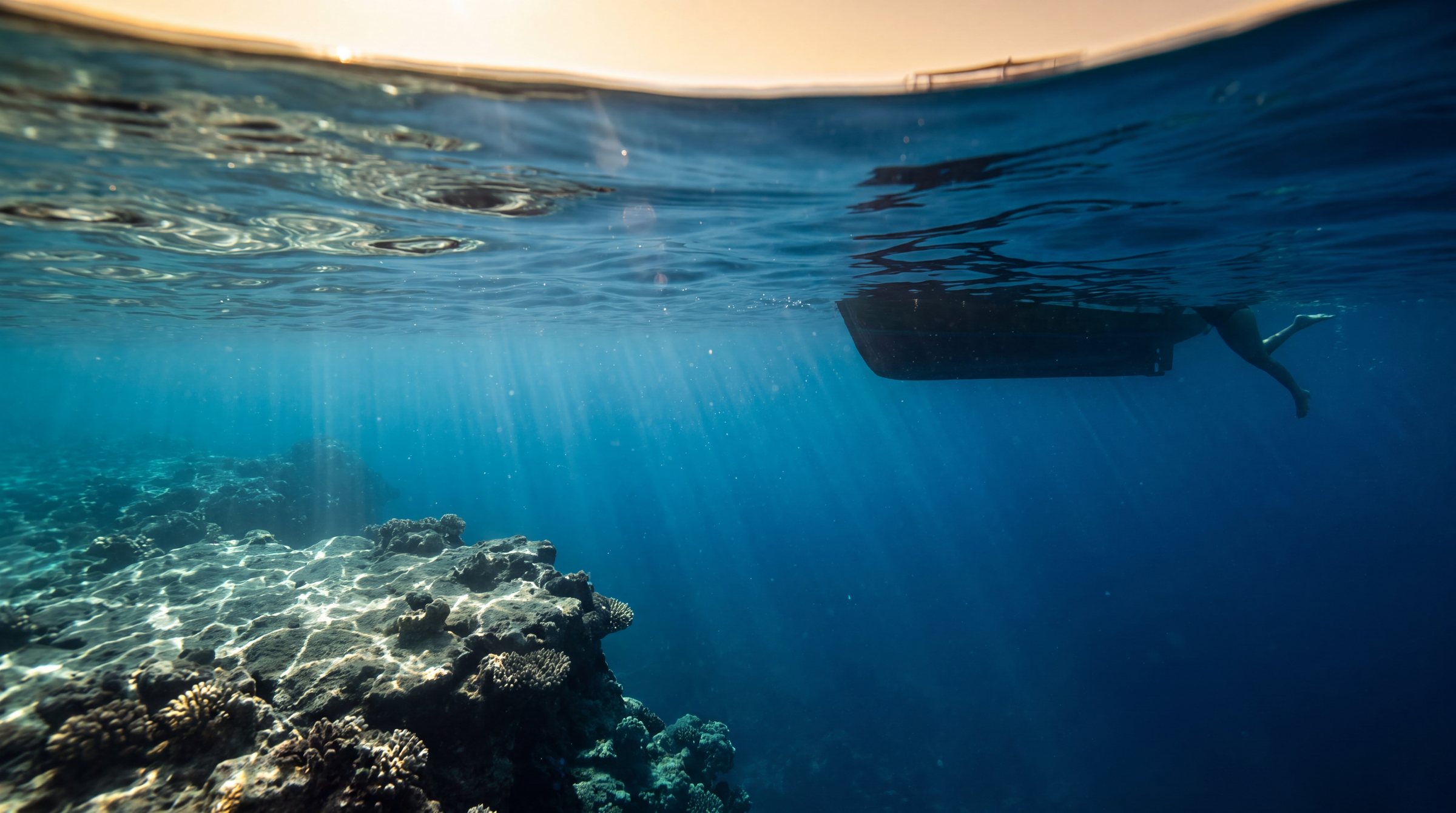 Net onder het oppervlak van de Rode Zee: zonlicht dat in banen door turkoois water op een koraalrichel valt, en de donkere romp van een bootje met een zwemmer ernaast.